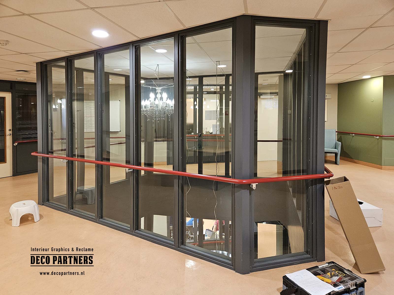 Glazen trappenhuis in zorginstelling vóór bestickering met zicht op trap en verdieping, met transparante glazen wanden en een open, onrustige uitstraling