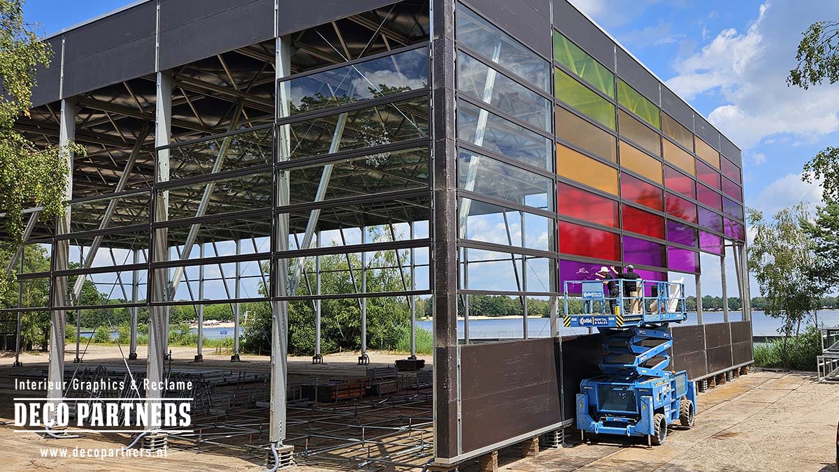 Monteurs brengen met een hoogwerker transparante gekleurde folie aan op de hoge glazen gevel van de Awakenings festivaltent