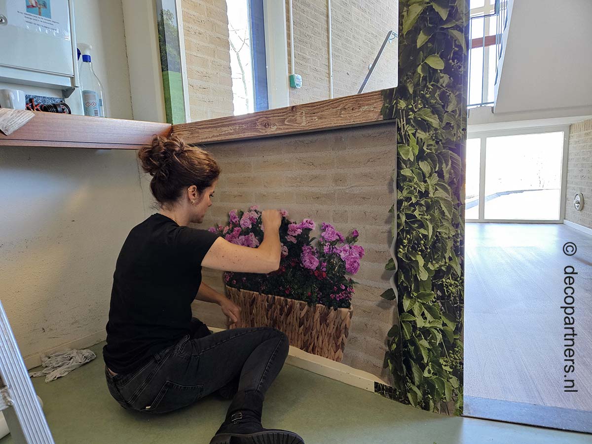 Medewerker van DecoPartners plakt muurprint met bloemenmand tijdens montage in verpleeghuis als onderdeel van dementievriendelijke inrichting.