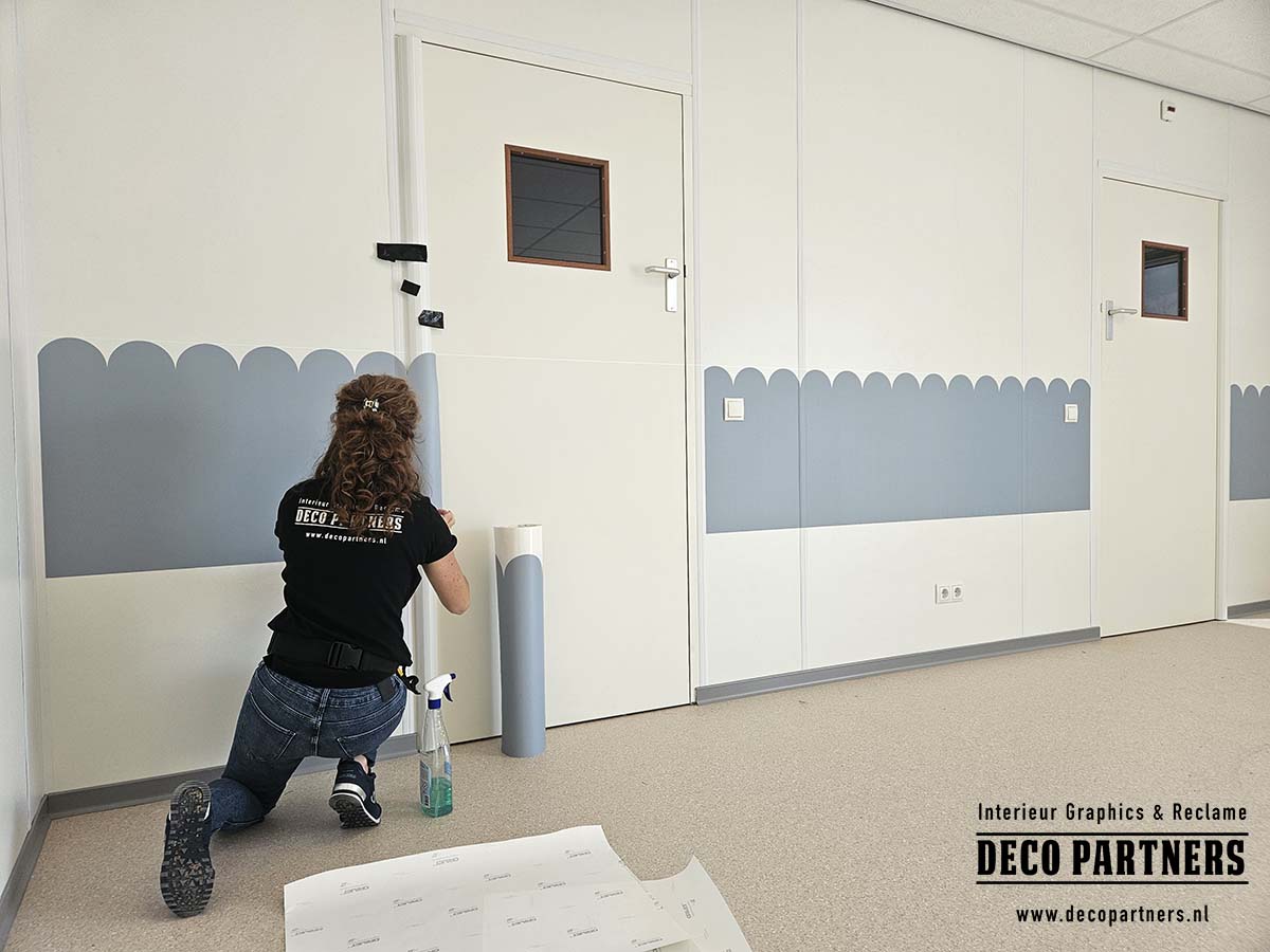 Monteur brengt lichtblauwe verwijderbare wandfolie met een uitgesneden schulprand aan bij de deuren van een kinderopvang.